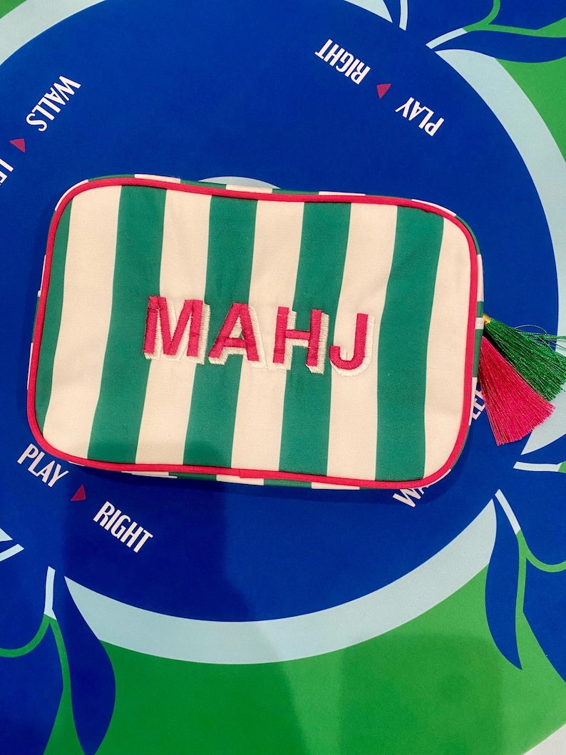 May include: A rectangular, striped pouch with green and white vertical stripes. The word "MAHJ" is embroidered in pink. The pouch has a red trim and a tassel with green and pink threads. The background is a blue and green game board.