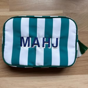 May include: Rectangular cosmetic bag with teal and white vertical stripes. The bag has the embroidered letters "MAHJ" in navy blue. A green tassel hangs from the side. The bag is on a wooden surface.