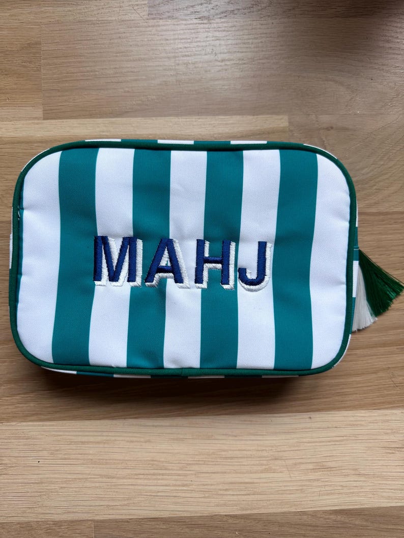 May include: Rectangular cosmetic bag with teal and white vertical stripes. The bag has the embroidered letters "MAHJ" in navy blue. A green tassel hangs from the side. The bag is on a wooden surface.
