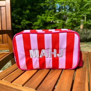 May include: A rectangular cosmetic bag with red and light pink vertical stripes. The bag has a pink zipper and trim, with the embroidered letters "MAHJ" in white. A small pink tassel hangs from the side. The bag is on a wooden surface.