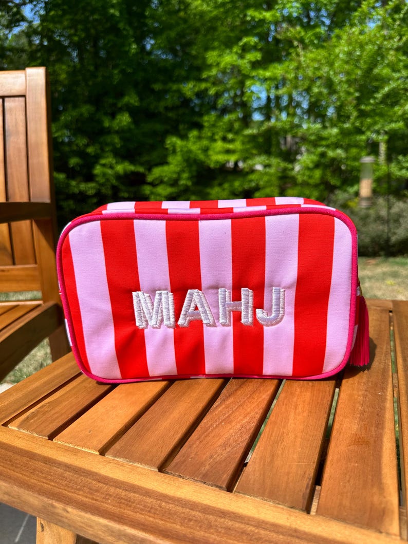 May include: A rectangular cosmetic bag with red and light pink vertical stripes. The bag has a pink zipper and trim, with the embroidered letters "MAHJ" in white. A small pink tassel hangs from the side. The bag is on a wooden surface.