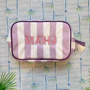 May include: A rectangular cosmetic bag with vertical white and lavender stripes. The bag features the embroidered letters "MAHJ" in pink and lavender. It has a purple zipper and trim, with a tassel on the side. The background has a palm tree pattern.