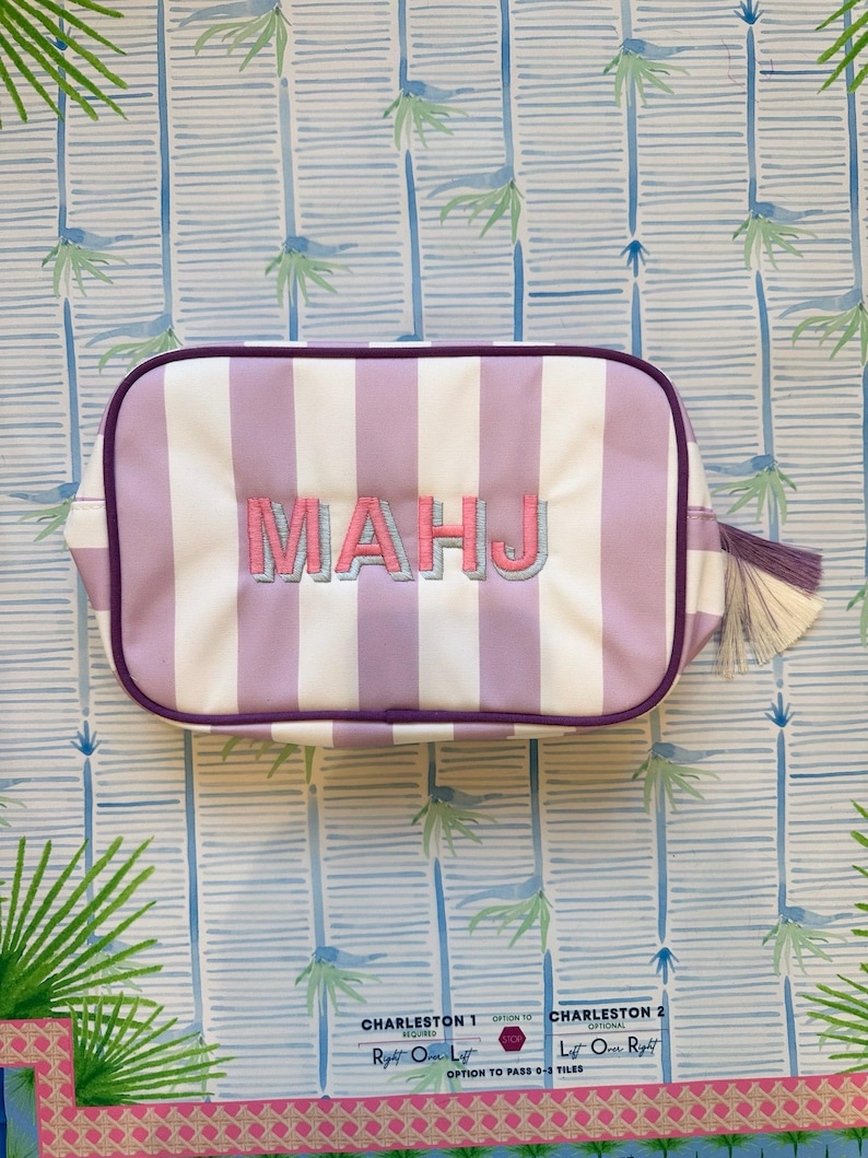 May include: A rectangular cosmetic bag with vertical white and lavender stripes. The bag features the embroidered letters "MAHJ" in pink and lavender. It has a purple zipper and trim, with a tassel on the side. The background has a palm tree pattern.