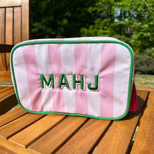 May include: A rectangular cosmetic bag with pink and white vertical stripes. The bag has green trim and the embroidered letters "MAHJ" in green. A pink tassel hangs from the side. The bag is sitting on a wooden surface.