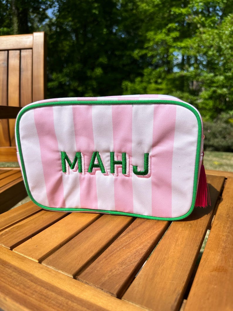 May include: A rectangular cosmetic bag with pink and white vertical stripes. The bag has green trim and the embroidered letters "MAHJ" in green. A pink tassel hangs from the side. The bag is sitting on a wooden surface.