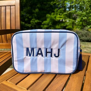 May include: A rectangular cosmetic bag with vertical stripes in light blue and white. The bag features the embroidered letters "MAHJ" in navy blue. A navy blue tassel hangs from the side. The bag is on a wooden table.