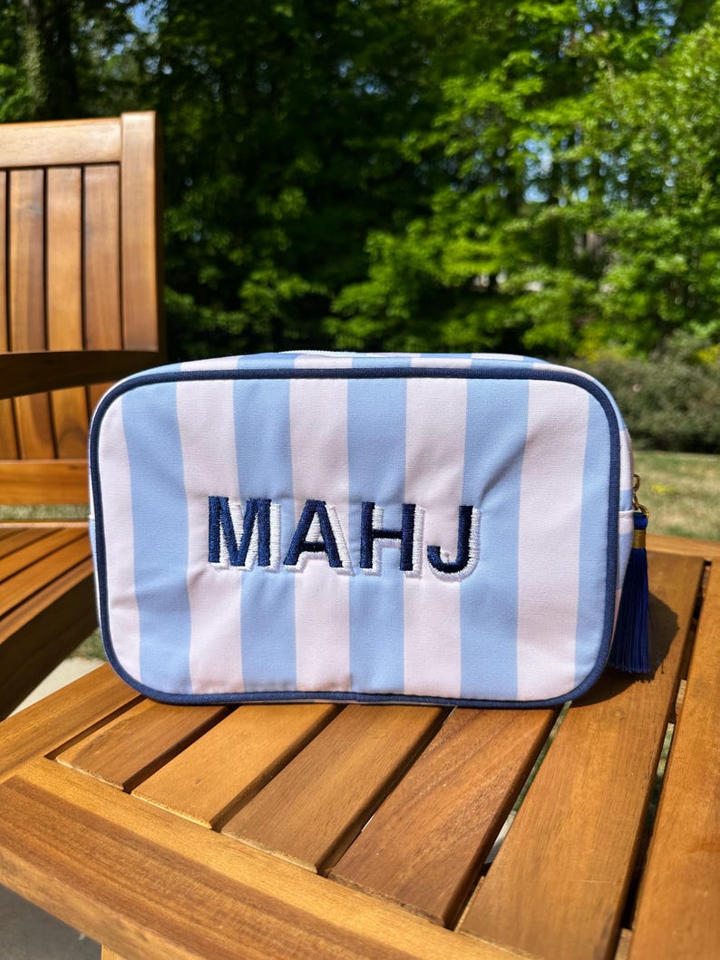 May include: A rectangular cosmetic bag with vertical stripes in light blue and white. The bag features the embroidered letters "MAHJ" in navy blue. A navy blue tassel hangs from the side. The bag is on a wooden table.