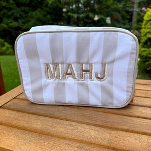 May include: A rectangular cosmetic bag with vertical beige and white stripes. The embroidered letters "MAHJ" are centered on the front. The bag is sitting on a wooden table, with a green background.