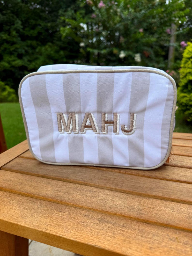May include: A rectangular cosmetic bag with vertical beige and white stripes. The embroidered letters "MAHJ" are centered on the front. The bag is sitting on a wooden table, with a green background.