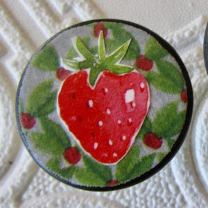 May include: Two circular cabinet knobs featuring a vibrant red strawberry illustration centered on a background of green leaves and small red berries. Each knob has a smooth, rounded black edge and a flat top surface with a detailed fruit graphic.