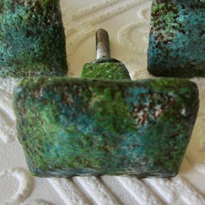 May include: Three square cabinet knobs with a textured, weathered surface featuring a mottled blend of deep green, teal, and dark brown tones. Each knob has a central metal mounting screw extending from the back for attachment to furniture.