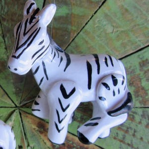 May include: Two ceramic zebra figurines. Each zebra is white with black stripes and markings. The figurines are posed in a seated position. The background is a green and brown wooden surface.