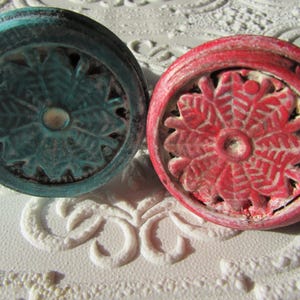 Puede incluir: Dos pomos redondos para armarios, uno con acabado en azul cerceta desgastado y otro en rojo desgastado. Cada pomo presenta un patrón floral o de copo de nieve en relieve en la cara frontal con una hendidura circular lisa en el centro.