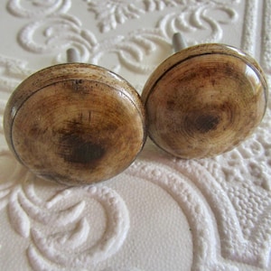 May include: Two round cabinet knobs featuring a polished, light brown wood grain finish with darker circular rings and a smooth, glossy surface. Each knob has a central metal mounting post extending from the back, designed for furniture attachment.