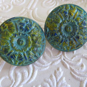 May include: Two round buttons featuring a raised, intricate floral pattern in shades of teal, lime green, and dark blue. The surface of each button has a textured, weathered finish with a central circular motif and radiating petal details.