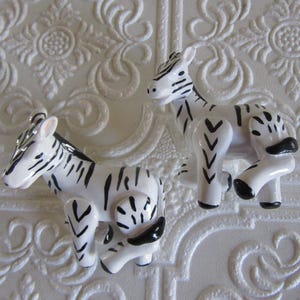 May include: Two small, glossy ceramic zebra figurines featuring a white base coat with hand-painted black stripes. Each zebra is depicted in a standing position with a smooth, reflective finish, showcasing detailed black ears, hooves, and tails.