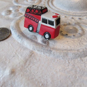 Pu&ograve; includere: Un piccolo camion dei pompieri rosso e bianco con dettagli neri, tra cui una scala e ruote. Il giocattolo &egrave; posto su una superficie bianca strutturata. C'&egrave; una moneta accanto.