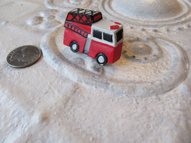 Pu&ograve; includere: Un piccolo camion dei pompieri rosso e bianco con dettagli neri, tra cui una scala e ruote. Il giocattolo &egrave; posto su una superficie bianca strutturata. C'&egrave; una moneta accanto.