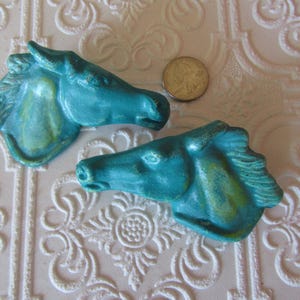May include: Two turquoise horse head sculptures with a metallic finish. The horse heads are side-facing, with detailed manes and facial features. A US quarter coin is next to the sculptures. The background is a white, textured surface.
