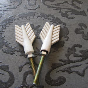 May include: Two white arrow-shaped cabinet knobs with a textured design. The knobs are attached to gold-colored screws with silver nuts. The background is a dark gray surface with a decorative pattern.