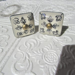 May include: Two square cabinet knobs with a cream background, black leaf design, and a gold-colored center. The knobs are likely made of ceramic or porcelain and are suitable for furniture or decor.