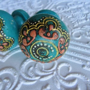 Puede incluir: Primer plano de dos pomos de cerámica turquesa con intrincados diseños pintados a mano. Los pomos presentan patrones naranjas, amarillos y negros sobre un fondo verde azulado. Los pomos son probablemente para muebles o uso decorativo.