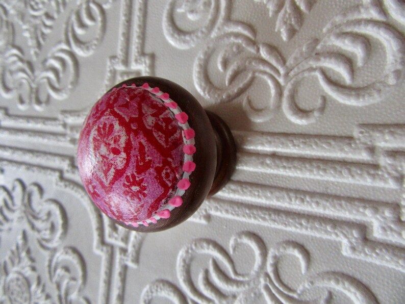 K&ouml;nnte beinhalten: Dekorativer Holzschrankknauf mit dunkelbraunem Sockel und rundem, gemustertem Oberteil. Die Oberseite zeigt ein rotes und rosa Blumenmuster mit einem Rand aus kleinen rosa Punkten. Der Knauf ist vor einem wei&szlig;en, strukturierten Hintergrund angebracht.