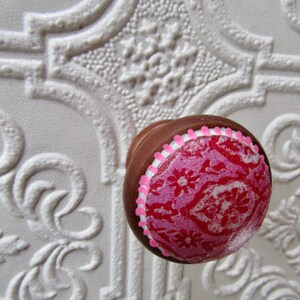 K&ouml;nnte beinhalten: Ein brauner Schrankknopf mit einem rosa-roten Blumenmuster. Der Knopf hat einen rosa-wei&szlig;en, wellenf&ouml;rmigen Rand. Der Hintergrund ist eine wei&szlig;e, strukturierte Oberfl&auml;che mit einem sich wiederholenden Muster.