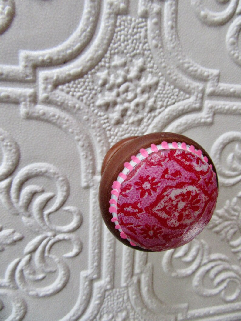 K&ouml;nnte beinhalten: Ein brauner Schrankknopf mit einem rosa-roten Blumenmuster. Der Knopf hat einen rosa-wei&szlig;en, wellenf&ouml;rmigen Rand. Der Hintergrund ist eine wei&szlig;e, strukturierte Oberfl&auml;che mit einem sich wiederholenden Muster.
