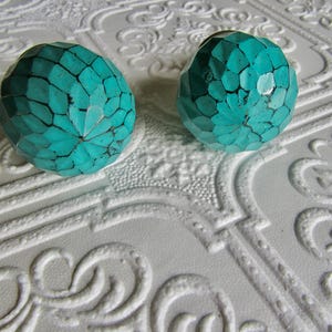 May include: Two turquoise cabinet knobs with a geometric design. The knobs are a vibrant teal color with dark lines separating the facets. They are set against a white, textured background with a repeating pattern.