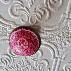K&ouml;nnte beinhalten: Ein runder, dekorativer Schrankknopf mit braunem Rand und einem rosa-roten Blumenmuster. Der Knopf hat einen rosa gepunkteten Rand und ist vor einem wei&szlig;en, strukturierten Hintergrund platziert.