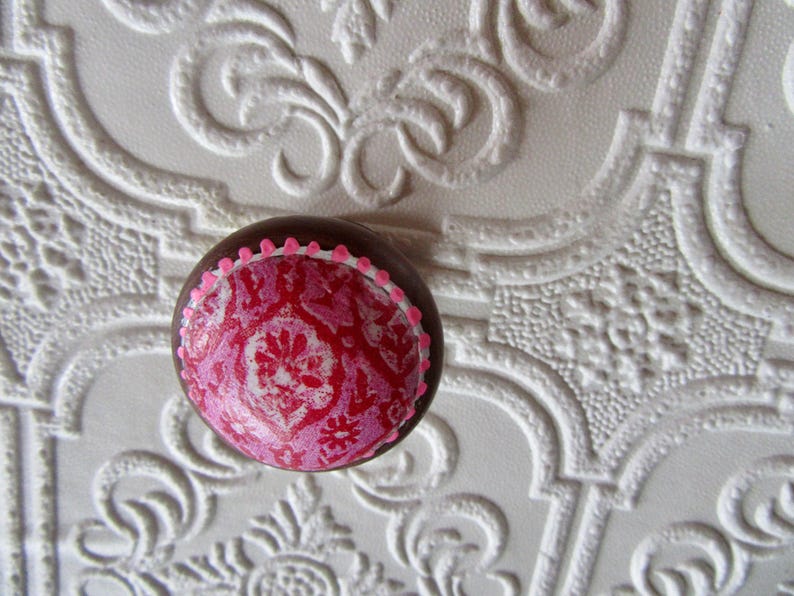 K&ouml;nnte beinhalten: Ein runder, dekorativer Schrankknopf mit braunem Rand und einem rosa-roten Blumenmuster. Der Knopf hat einen rosa gepunkteten Rand und ist vor einem wei&szlig;en, strukturierten Hintergrund platziert.