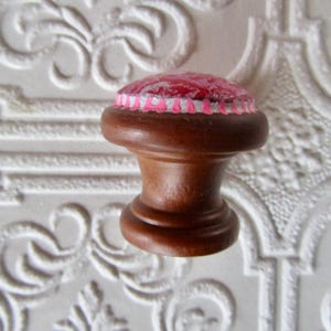 K&ouml;nnte beinhalten: Dekorativer Schrankknopf aus Holz mit dunkelbraunem Finish. Der Knopf hat eine runde Oberseite mit einem rosa und roten Design, akzentuiert mit wei&szlig;en und rosa Zierleisten. F&uuml;r M&ouml;bel und Heimdekoration.