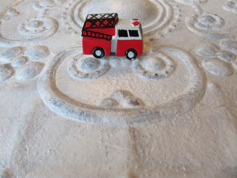 Pu&ograve; includere: Un piccolo camion dei pompieri rosso e bianco con ruote nere e una scala sulla parte superiore. Il giocattolo &egrave; posto su uno sfondo bianco strutturato con motivi ornamentali.