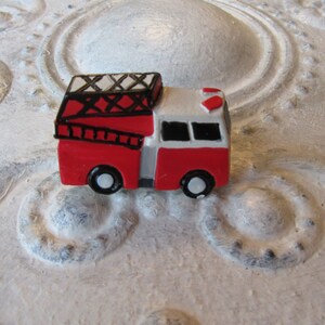 Pu&ograve; includere: Una statuetta di un camion dei pompieri rosso e bianco in miniatura con ruote nere e una scala. Il camion dei pompieri giocattolo ha un tetto nero con un design bianco e nero. La statuetta &egrave; ambientata su uno sfondo strutturato bianco sporco.