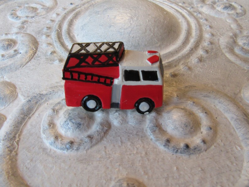Pu&ograve; includere: Una statuetta di un camion dei pompieri rosso e bianco in miniatura con ruote nere e una scala. Il camion dei pompieri giocattolo ha un tetto nero con un design bianco e nero. La statuetta &egrave; ambientata su uno sfondo strutturato bianco sporco.