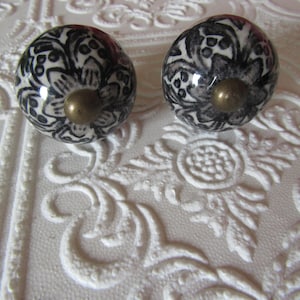May include: Two ceramic cabinet knobs with a black and white floral design. Each knob has a brass-colored metal base. The knobs are round and sit on a white surface with a raised floral pattern.