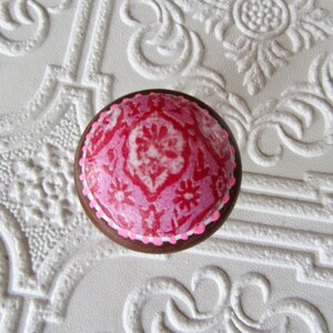 K&ouml;nnte beinhalten: Runder, dekorativer Schrankknopf mit einem rosa-wei&szlig;en Blumenmuster auf dunkelbraunem Grund. Der Knopf hat einen wei&szlig;en Rand und ist vor einem strukturierten wei&szlig;en Hintergrund platziert.