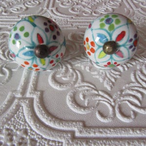 May include: Two white ceramic cabinet knobs with colorful floral designs. Each knob features a pattern of teal, red, orange, yellow, green, and purple dots and petal shapes. The knobs have bronze-colored metal hardware.