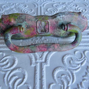 May include: A distressed metal drawer pull with a keyhole, painted in shades of pink, green, and brown. The handle is curved and attached to a decorative backplate. This vintage-style hardware adds a unique touch to furniture.