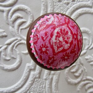 K&ouml;nnte beinhalten: Ein runder Holzknauf mit einem rosa-roten Blumenmuster. Der Knauf hat einen braunen Rand und ist mit kleinen rosa Punkten verziert. Der Hintergrund ist eine wei&szlig;e, strukturierte Oberfl&auml;che.