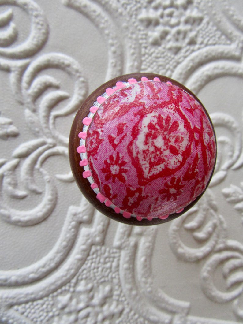 K&ouml;nnte beinhalten: Ein runder Holzknauf mit einem rosa-roten Blumenmuster. Der Knauf hat einen braunen Rand und ist mit kleinen rosa Punkten verziert. Der Hintergrund ist eine wei&szlig;e, strukturierte Oberfl&auml;che.
