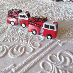 Pu&ograve; includere: Due camion dei pompieri rossi e bianchi in miniatura con dettagli neri e scale. I giocattoli sono su una superficie bianca strutturata con un motivo floreale ripetuto. I camion hanno ruote nere.