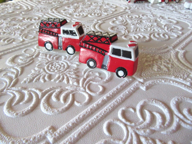 Pu&ograve; includere: Due camion dei pompieri rossi e bianchi in miniatura con dettagli neri e scale. I giocattoli sono su una superficie bianca strutturata con un motivo floreale ripetuto. I camion hanno ruote nere.