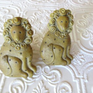 May include: Two ceramic lion figurines with curly manes and spotted bodies. The lions are sitting, with their paws resting on their legs. The figurines are a muted yellow-green color and have small black dots. The background is a white, textured surface.
