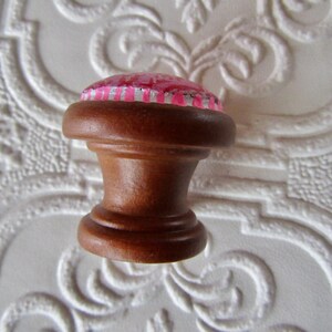 K&ouml;nnte beinhalten: Ein brauner Holzschrankknopf mit einem rosa und silber gemusterten Oberteil. Der Knopf hat ein klassisches, gedrehtes Design und wird wahrscheinlich f&uuml;r M&ouml;bel oder dekorative Zwecke verwendet. Der Knopf ist vor einem wei&szlig;en Hintergrund platziert.