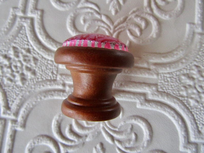 K&ouml;nnte beinhalten: Ein brauner Holzschrankknopf mit einem rosa und silber gemusterten Oberteil. Der Knopf hat ein klassisches, gedrehtes Design und wird wahrscheinlich f&uuml;r M&ouml;bel oder dekorative Zwecke verwendet. Der Knopf ist vor einem wei&szlig;en Hintergrund platziert.