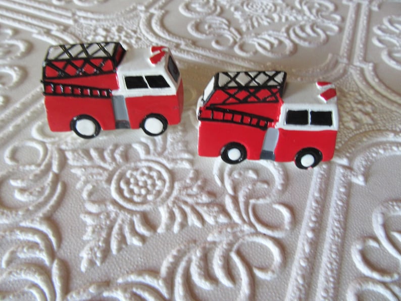 May include: Two miniature red and white fire trucks with black detailing and wheels. The trucks have ladders and a roof rack. The background is a white, textured surface with a floral pattern.