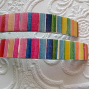 May include: Two colorful hair barrettes with a striped pattern in shades of pink, blue, yellow, and green. The barrettes are curved and appear to be made of metal or a similar material. The background is a textured white surface.