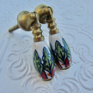 May include: Two vintage brass door handles with white ceramic grips. The ceramic grips are hand-painted with a design of green leaves, blue accents, and red details. The handles are set against a textured white surface.